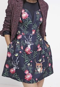 Sininen kukkakuosiset mekko, jossa on hirvi ja lintuja, ja joka sisältää istuva yläosa, pyöreä pääntie, suuret taskut ja levenevä hame.