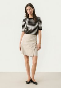 Femme portant un haut à rayures noires et blanches à manches courtes, une jupe portefeuille beige avec un bouton et des ballerines noires, se tenant sur un fond neutre.