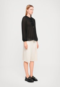 Svart transparent bluse med volanger foran og ved ermene, kombinert med lysebeige knelengde shorts. Svart loafers med detaljer i gull.