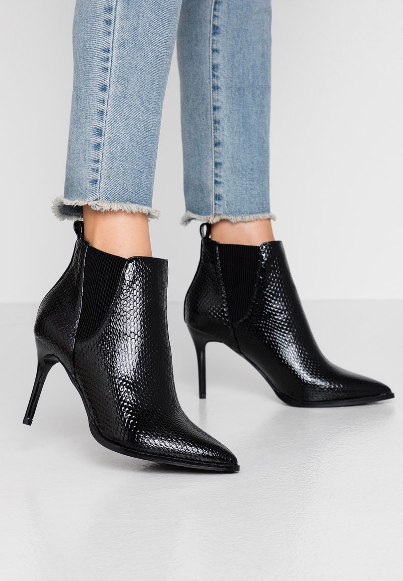 Sorte ankelstøvler i slangeskindslook med spidse tæer, stilethæle og elastiske sidelommer, kombineret med slidte blå jeans.
