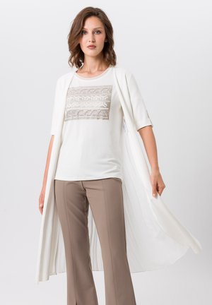 Haut blanc avec une broderie beige complexe, associé à un pantalon flare beige. La tenue est complétée par un long cardigan blanc à superpositions.
