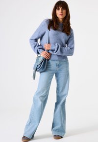 Eine Frau trägt einen hellblauen, perlenbesetzten Pullover, weite Jeans, graue Schuhe und hält eine blaue Umhängetasche mit Quaste. Sie steht vor einem weißen Hintergrund.