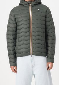 Veste matelassée verte avec capuche et fermeture éclair multicolore, portée avec un jean bleu clair.