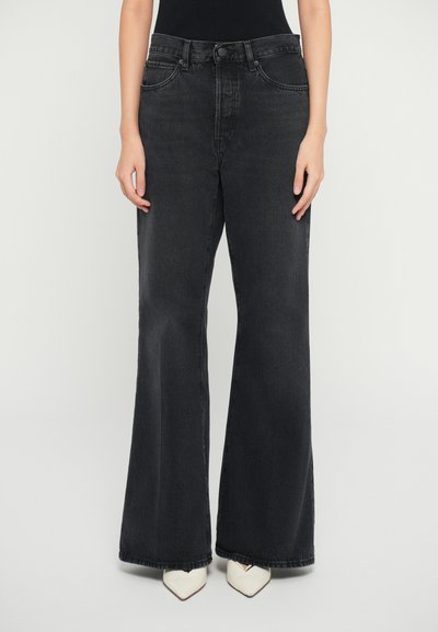 Jean large noir en denim, avec une taille haute, un design à cinq poches et des détails cousus. Associé à des talons blancs pointus.