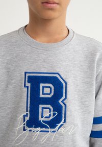 Szary sweatshirt z niebieską łatą z literą "B" i białym obramowaniem. Posiada niebieskie paski na rękawach. Miękka tekstura i fason z okrągłym dekoltem.