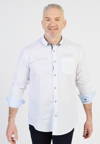 Chemise blanche à petits pois multicolores, col boutonné et poignets intérieurs bleus contrastants. Comprend une poche latérale avec un détail de logo.