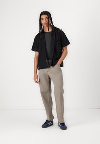 Black short-sleeve button-up shirt layered over a fitted gray tee, paired with loose light brown pants and blue sneakers with stripes.
