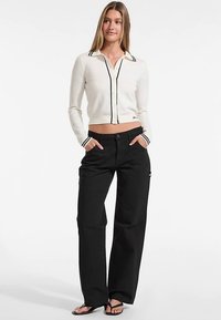 Weißer kurz geschnittener Cardigan mit schwarzen Akzenten, Knopfverschluss und spitzem Kragen; kombiniert mit schwarzen, weit geschnittenen Hosen und Sandalen.