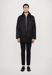 Black waterproof jacket with a button-up front, side pockets, and a relaxed fit, worn over a light-colored shirt and paired with slim trousers.