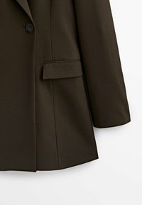 Blazer marron sur mesure avec une texture lisse, comprenant un seul bouton noir, une poche latérale et une coupe structurée.
