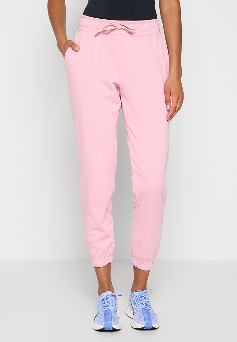 Osoba w jasnoróżowych spodniach dresowych z elastycznymi mankietami i niebieskich butach sportowych, stojąca z jedną ręką w kieszeni.