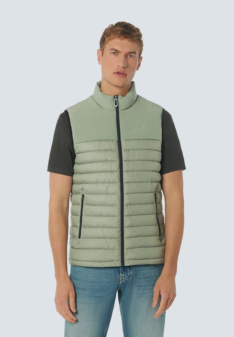 Gilet matelassé vert clair avec un col haut, fermeture éclair et deux poches latérales, porté par-dessus une chemise noire à manches courtes. Texture lisse.