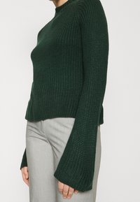 Pull en maille vert foncé avec un col montant, une texture côtelée et des manches évasées, associé à un pantalon ajusté gris clair.