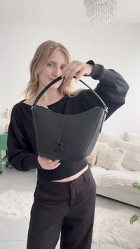 Νεαρή γυναίκα με ξανθά μαλλιά σε μαύρα ρούχα κρατά μια μαύρη τσάντα με μία λαβή σε ένα φωτεινό σαλόνι.
