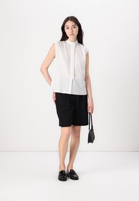 Weiße kurzärmlige Bluse aus strukturiertem Stoff mit Knöpfen vorne, kombiniert mit schwarzen knielangen Shorts und schwarzen Loafers. Kleine schwarze Handtasche.