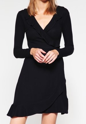 Femme portant une robe portefeuille noire à manches longues avec des détails à volants au niveau de l'encolure et de l'ourlet, posant devant un fond blanc uni.