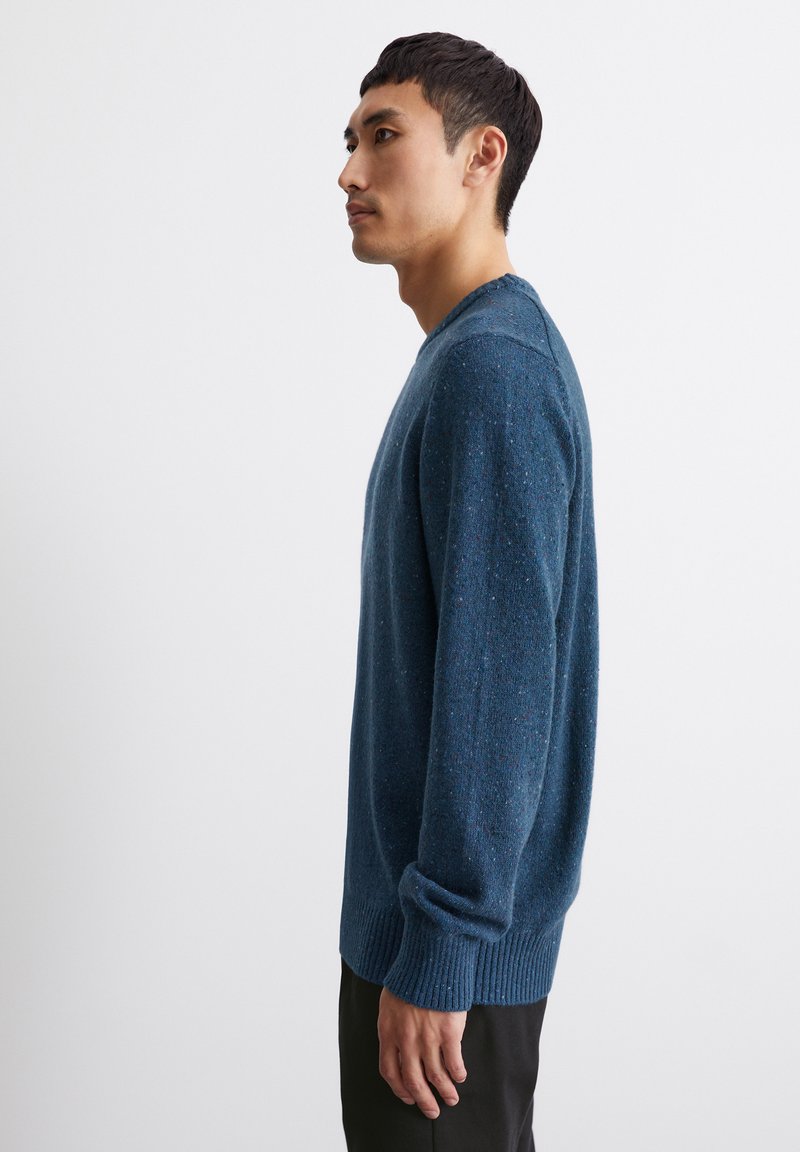 Ein Mann, der nach links schaut, trägt einen blauen melierten Strickpullover und schwarze Hosen vor einem schlichten weißen Hintergrund.