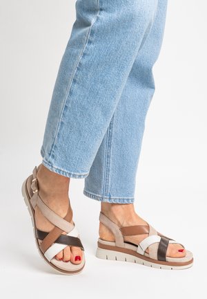Füße in Sandalen mit beige, braunen und weißen überkreuzten Riemen und dicken Sohlen, kombiniert mit hellblauen Jeans.