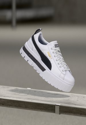 Bílá teniska Puma s černým pruhem a silnou podrážkou, zachycena ve vzduchu nad betonovou lavičkou na rozostřeném neutrálním pozadí.