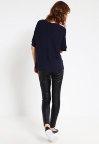 Nainen, jolla on hartioille ulottuvat hiukset, pukeutuneena tummansiniseen ribbikuvioituun lyhythihaiseen paitaan, mustiin nahkahousuihin ja mustiin liukuviin kenkiin, kävelee poispäin valkoista taustaa vasten.