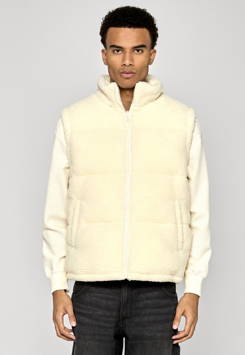 Jeune homme portant un gilet en polaire écru sur une chemise crème à manches longues et un jean noir, debout devant un fond gris uni.