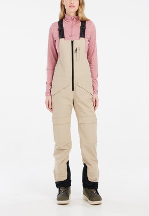 Beige Skianzüge mit verstellbaren Trägern, Frontreißverschluss und Cargo-Taschen. Schwarze Akzente an den Knöcheln. Das Modell trägt ein pinkes Langarmshirt.