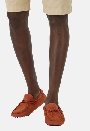 Orange suede loafers with a bow detail, featuring a rounded toe and rubber outsole, worn with khaki shorts.