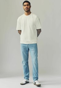 Tricou alb din bumbac, cu mânecă scurtă, asortat cu blugi drepti de culoare albastru deschis și adidași albi cu accente negre și detalii gri.