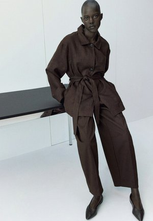 Persona con cabello corto que lleva un abrigo marrón oscuro con cinturón y pantalones de pierna ancha, apoyada en una mesa negra en una habitación blanca y minimalista.