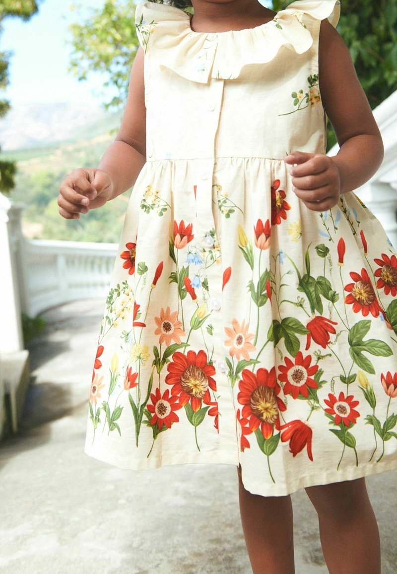 Tout-petit portant une robe crème sans manches avec un imprimé floral rouge et orange, debout sur une terrasse en pierre avec une rambarde blanche en extérieur.