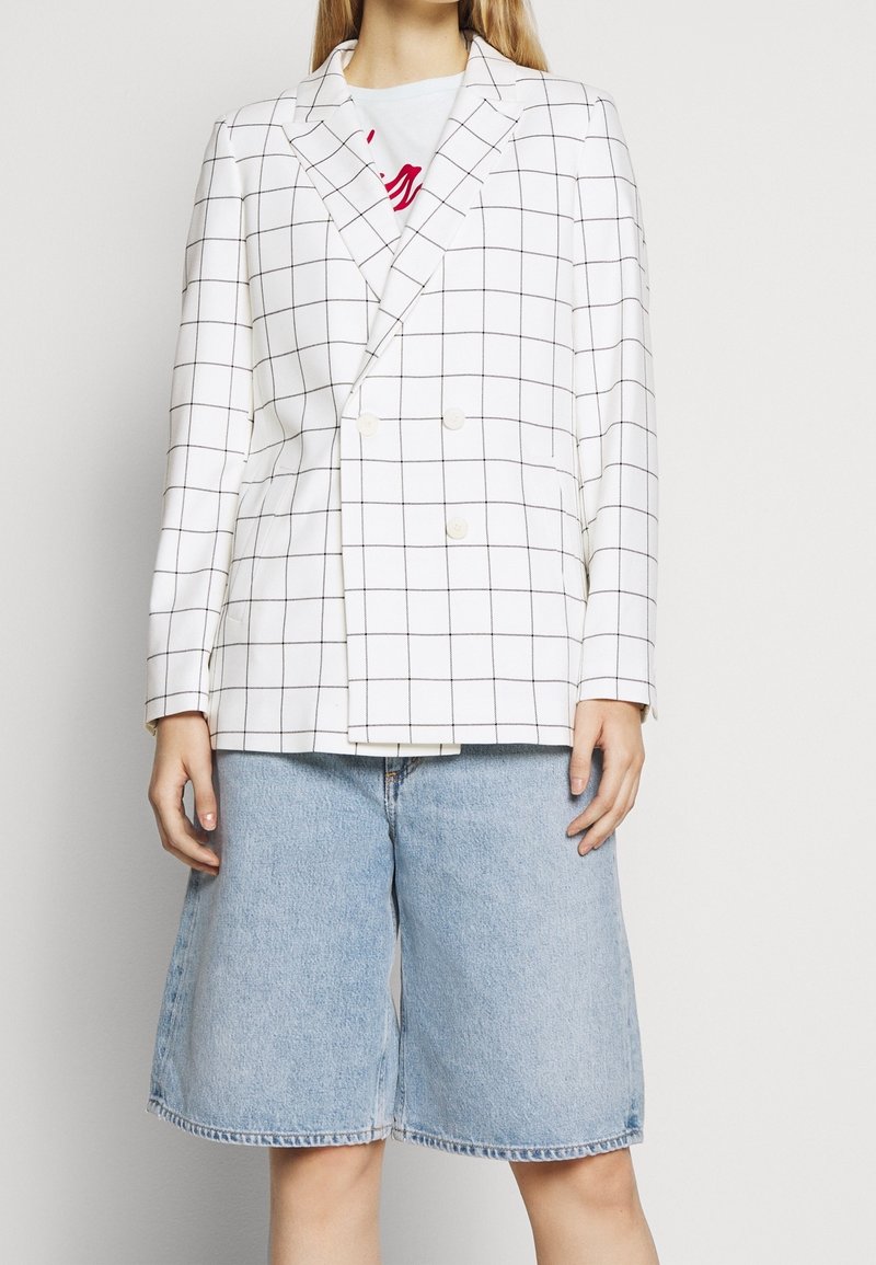 Blazer double boutonnage en tissu blanc avec un motif à carreaux noirs ; doté de revers crantés et de deux grands boutons. Associé à un short en denim bleu.