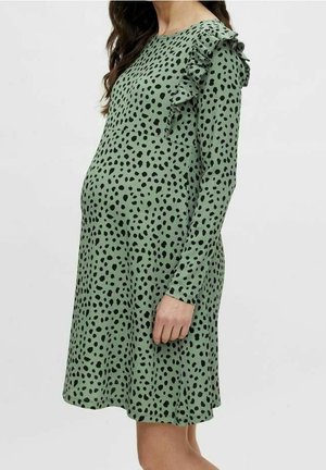 Vihreä mekko mustilla pilkuilla, pitkät hihat ja rypistetty yksityiskohta yhdessä olkapäässä, valmistettu pehmeästä ja kevyestä kankaasta.