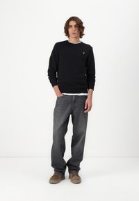 Zwarte sweatshirt met geribbelde hals, gecombineerd met losse grijze spijkerbroek en bruine schoenen. Opvallend goudkleurig logo op de borst.