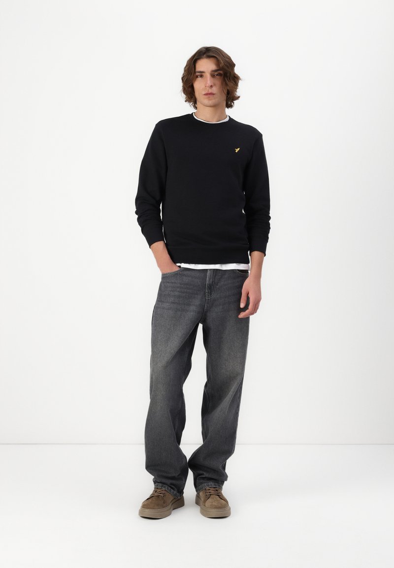Zwarte sweatshirt met geribbelde hals, gecombineerd met losse grijze spijkerbroek en bruine schoenen. Opvallend goudkleurig logo op de borst.