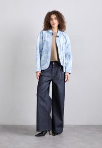 Veste bleu clair à motifs avec un col arrondi, portée sur un haut beige, associée à un pantalon en denim foncé à jambes larges et des chaussures noires à talons pointus.