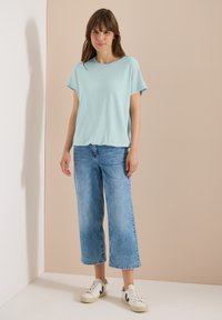 Femme debout contre un mur beige portant un haut bleu clair à manches courtes, un jean bleu large et court, et des baskets blanches avec des accents noirs.