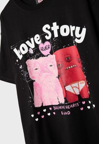 T-shirt noir avec deux monstres dessinés, un rose et un rouge, sous le texte « Love Story 4ever » et « Breaking Hearts & Wind » entouré de cœurs roses et blancs.