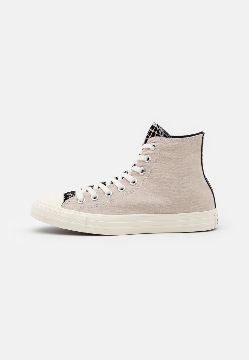 Basket montante en toile beige avec semelle en caoutchouc blanche et lacets, avec un motif texturé noir et blanc sur la languette et l'avant-pied.