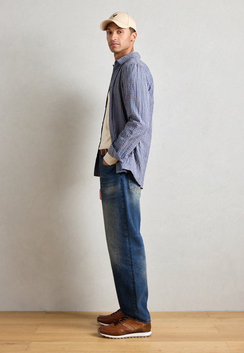 Tenue décontractée comprenant une chemise à carreaux bleue, une casquette beige, un t-shirt crème, un jean bleu délavé et des baskets marron avec des semelles blanches.