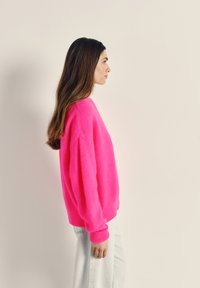 Sieviete ar gariem brūniem matiem, ģērbta spilgti rozā džemperī un gaišās džinsa biksēs, stāv profilā pret vienkāršu bēša krāsas fonu.