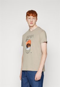 Beige katoenen T-shirt met NASA-afbeelding met tekst, oranje planeetillustratie en zwarte omlijning. Korte mouwen en ronde hals.