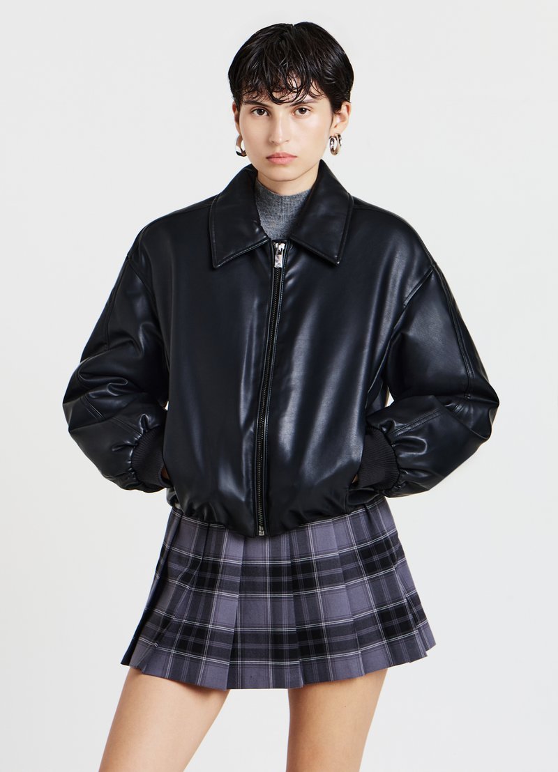 Giubbotto bomber in pelle sintetica nera con colletto ampio e chiusura con zip; abbinato a una gonna plissettata grigia a quadri con dettagli neri.