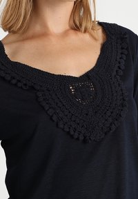 Marineblaues Shirt mit einem strukturierten, kunstvollen Häkelkragen mit gewelltem Rand und kleinen Bobbelakzenten. Weicher Stoff mit lockerer Passform.