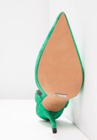 Chaussure à talon haut verte avec un bout pointu, tige en tissu texturé, semelle en cuir et détail de logo métallique au bas.