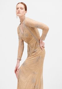 Beaded, naakt lange mouwen jurk met een V-hals, ingewikkelde zilveren kraal details in zonnestraalpatronen, en een rok met een hoge split tot boven de dij.