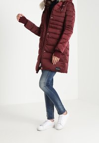 Person som bär en vinröd vadderad rock med pälshuva, blå jeans och vita sneakers, stående mot en enkel vit bakgrund.