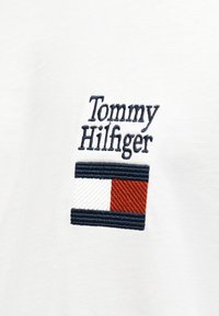 Λευκό ύφασμα με κεντημένο κείμενο "Tommy Hilfiger" πάνω από το ορθογώνιο λογότυπο της μάρκας σε σκούρο μπλε, λευκό και κόκκινο νήμα.