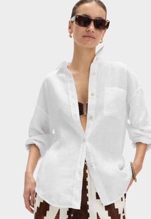Kvinde iført oversized hvid skjorte med knapper over sort bikinitop, brune og hvide mønstrede bukser, store solbriller og guldringøreringe.