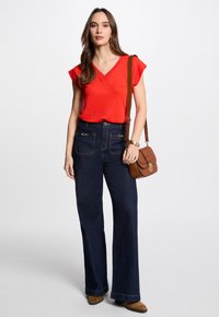 Rode V-hals shirt, blauwe wijde jeans met voorzakken, bruine schoudertas met schouderband, en bruine enkellaarsjes. Eenvoudig ontwerp en gladde textuur.