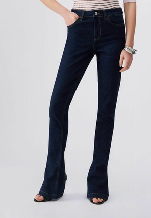 Donna con jeans bootcut blu scuro, top a righe, braccialetti d'argento, con una mano in tasca e scarpe nere aperte in punta visibili.